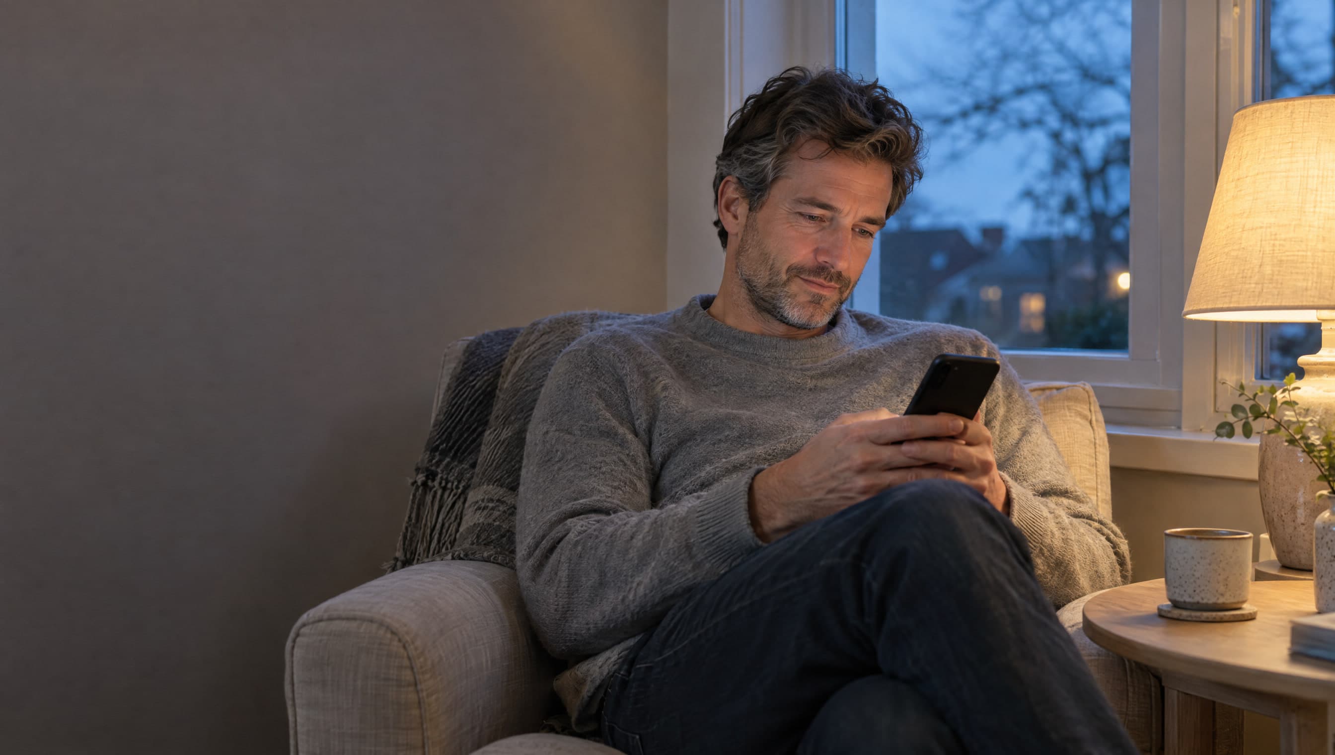 A man in a grey sweater in a linen armchair at dusk, reading on his phone beside a lit lamp, a cup on the side table.