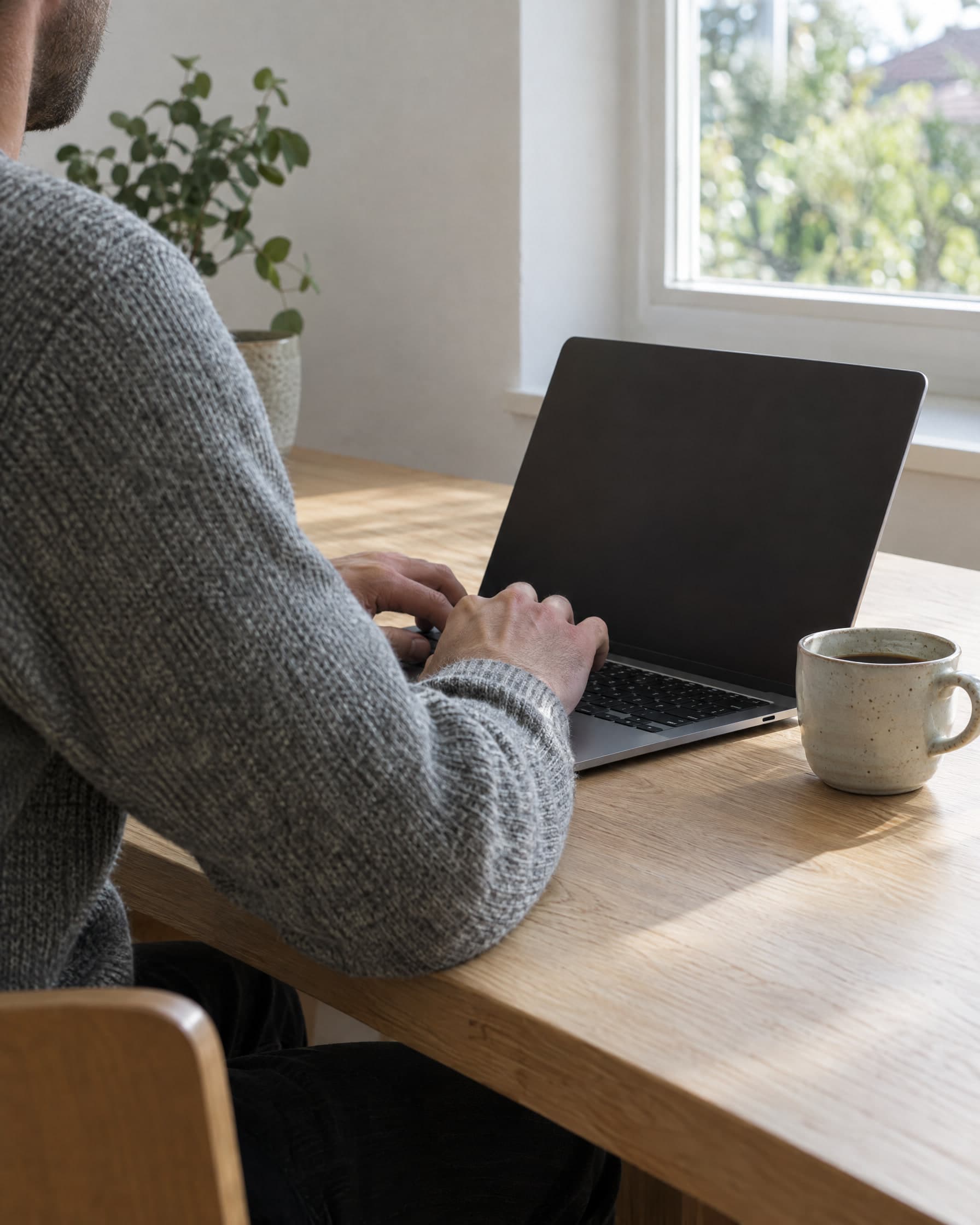 A man in a grey sweater at a kitchen table, typing on a laptop with a coffee cup beside it.