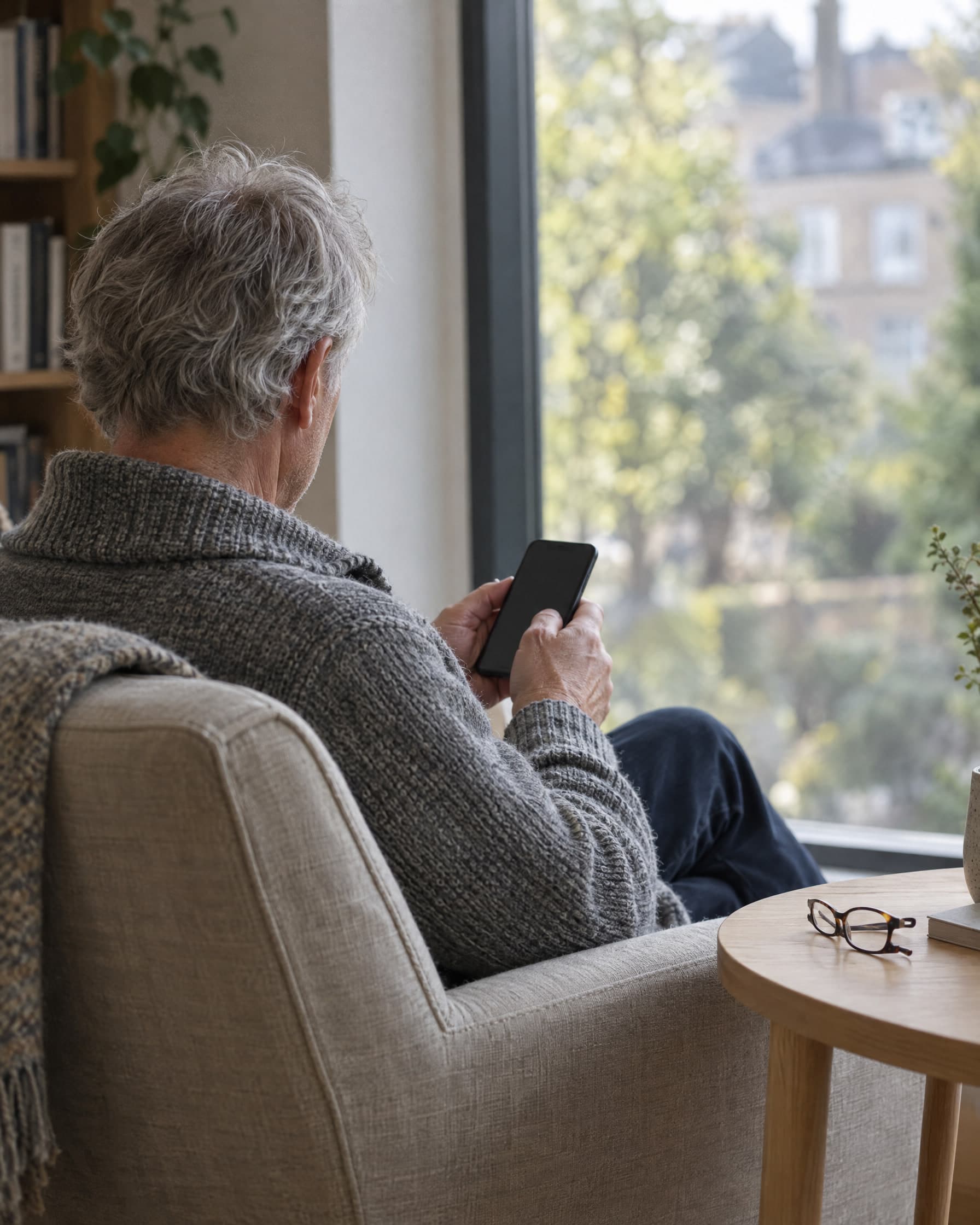 An older man reading on his phone in a linen armchair by a tall window, seen from behind.