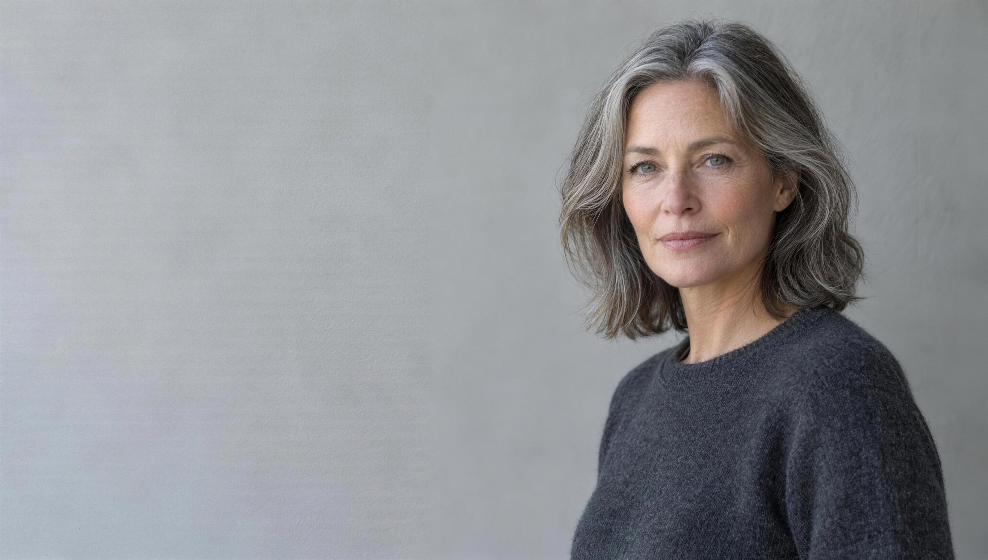 A woman in her early fifties with shoulder-length grey hair and a charcoal sweater, looking calmly at the camera against a pale grey wall.