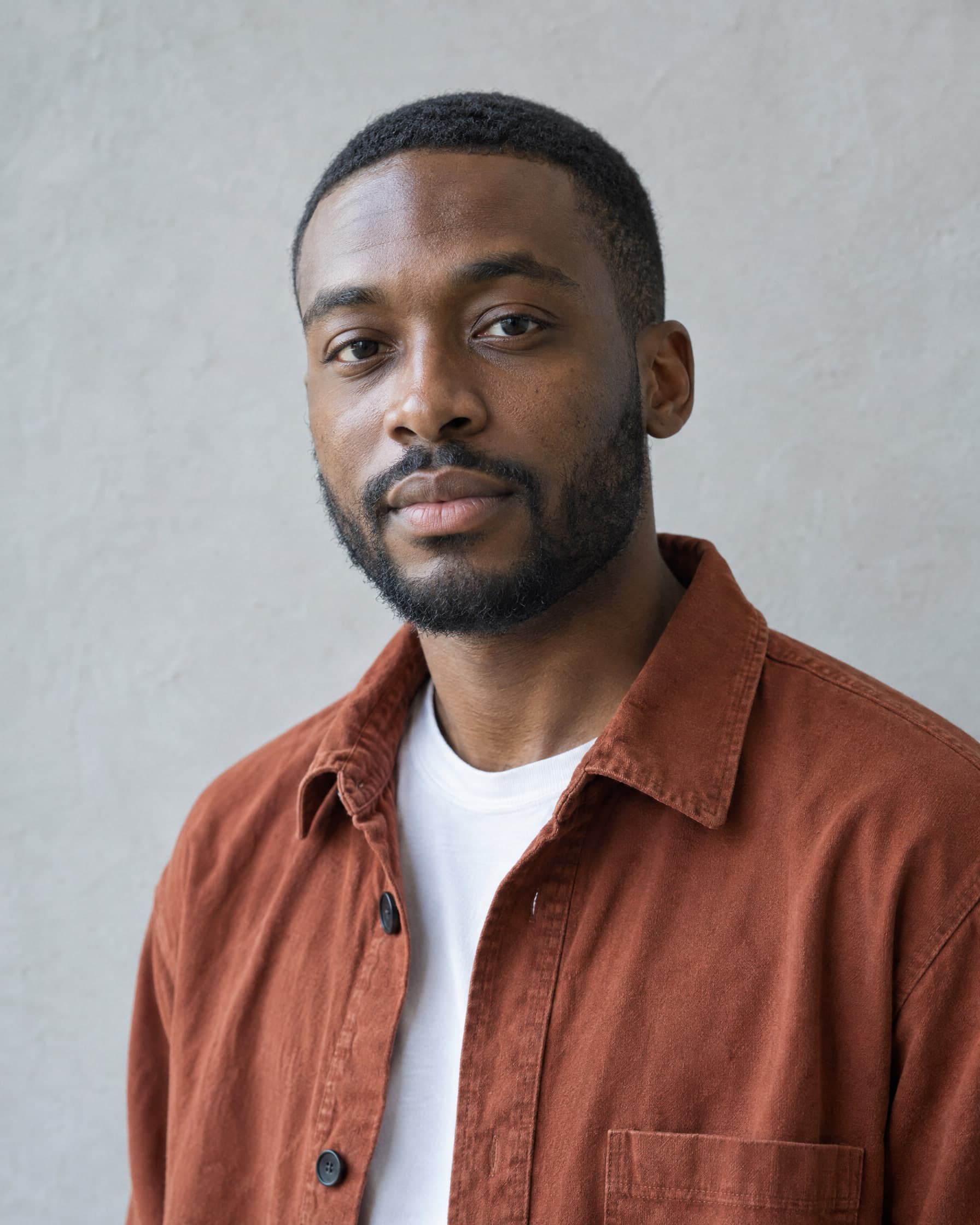 A man in his early thirties with a short beard, in a rust-coloured overshirt over a white T-shirt, looking calmly at the camera against a pale grey wall.