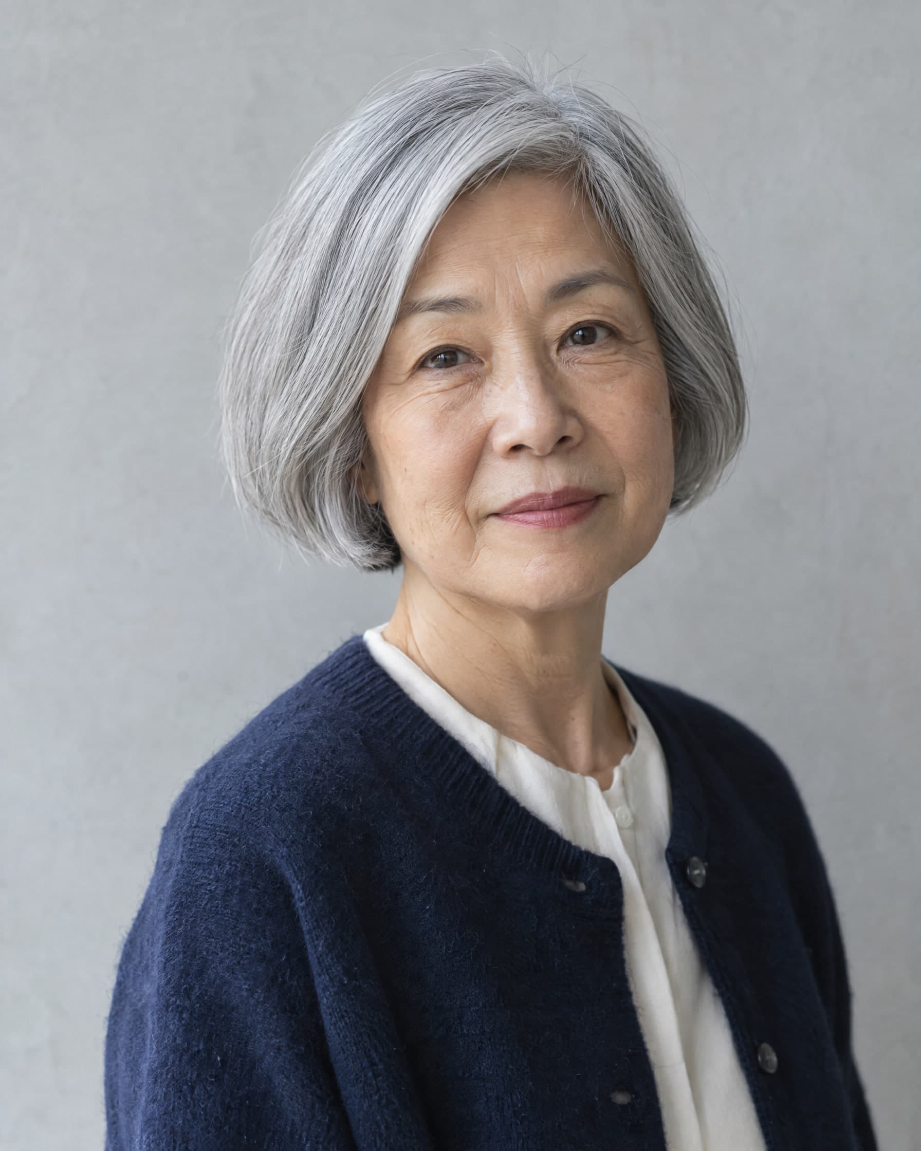 A woman in her late sixties with a silver bob, in a navy cardigan over a cream blouse, looking calmly at the camera against a pale grey wall.