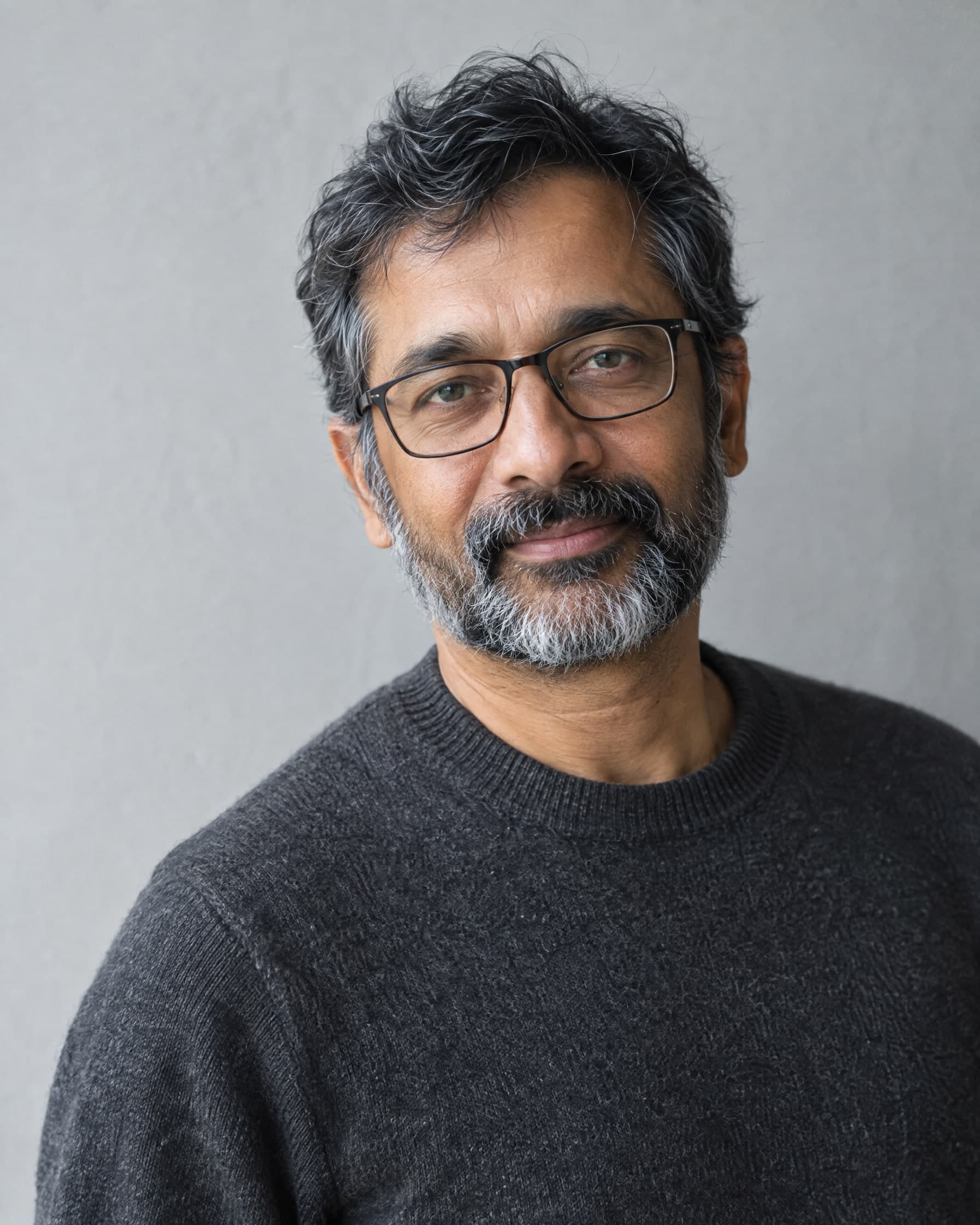A man in his mid-fifties with a salt-and-pepper beard and glasses, in a charcoal sweater, looking warmly at the camera against a pale grey wall.