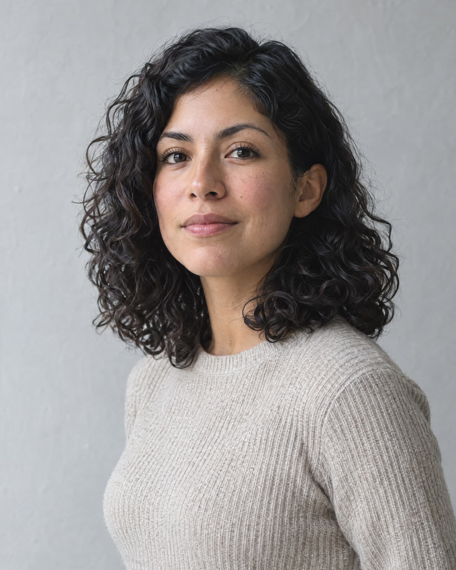 A woman in her late twenties with curly dark hair, in an oatmeal knit sweater, looking calmly at the camera against a pale grey wall.