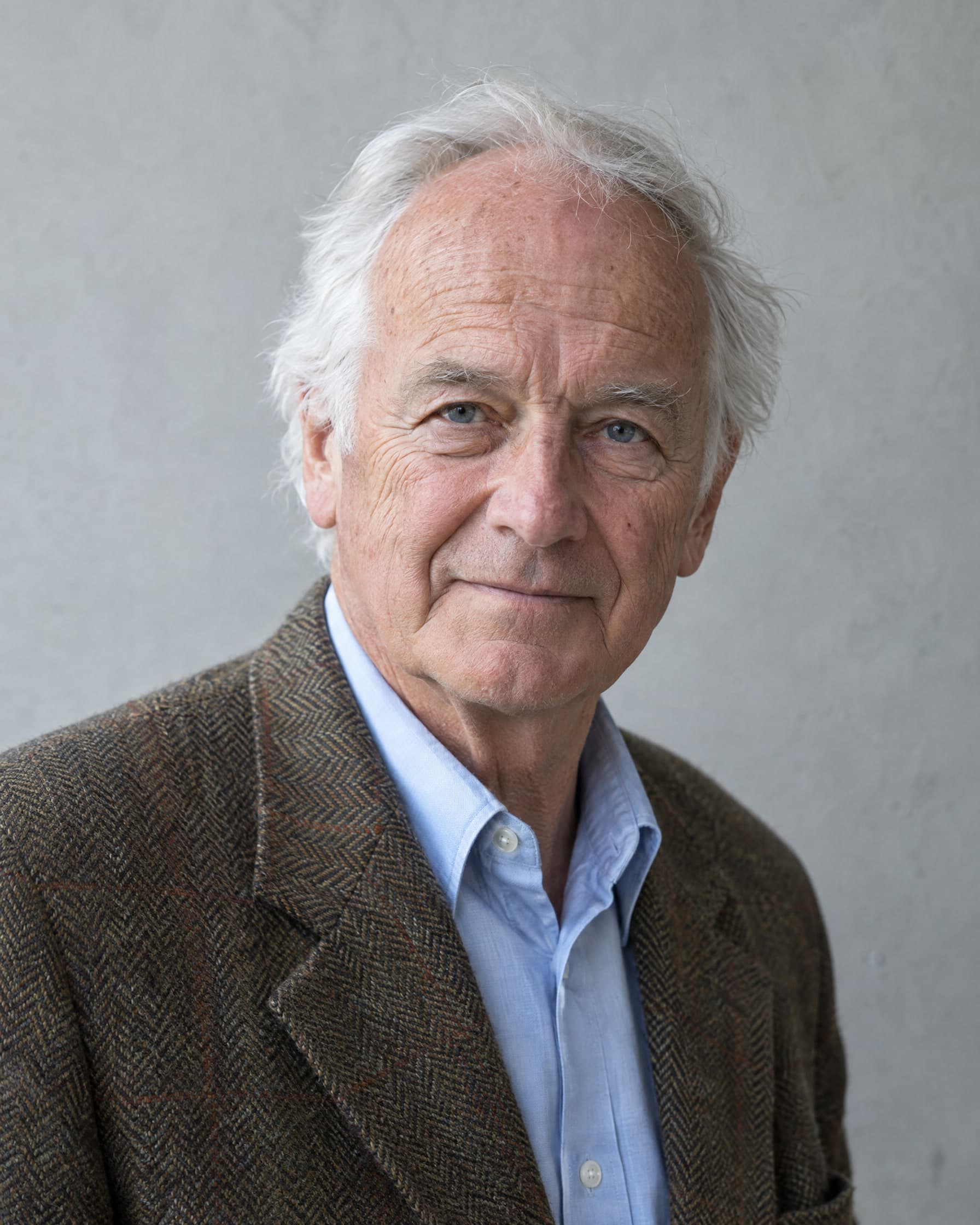A man in his early seventies with white hair, in a brown tweed jacket and a pale blue shirt, looking kindly at the camera against a pale grey wall.