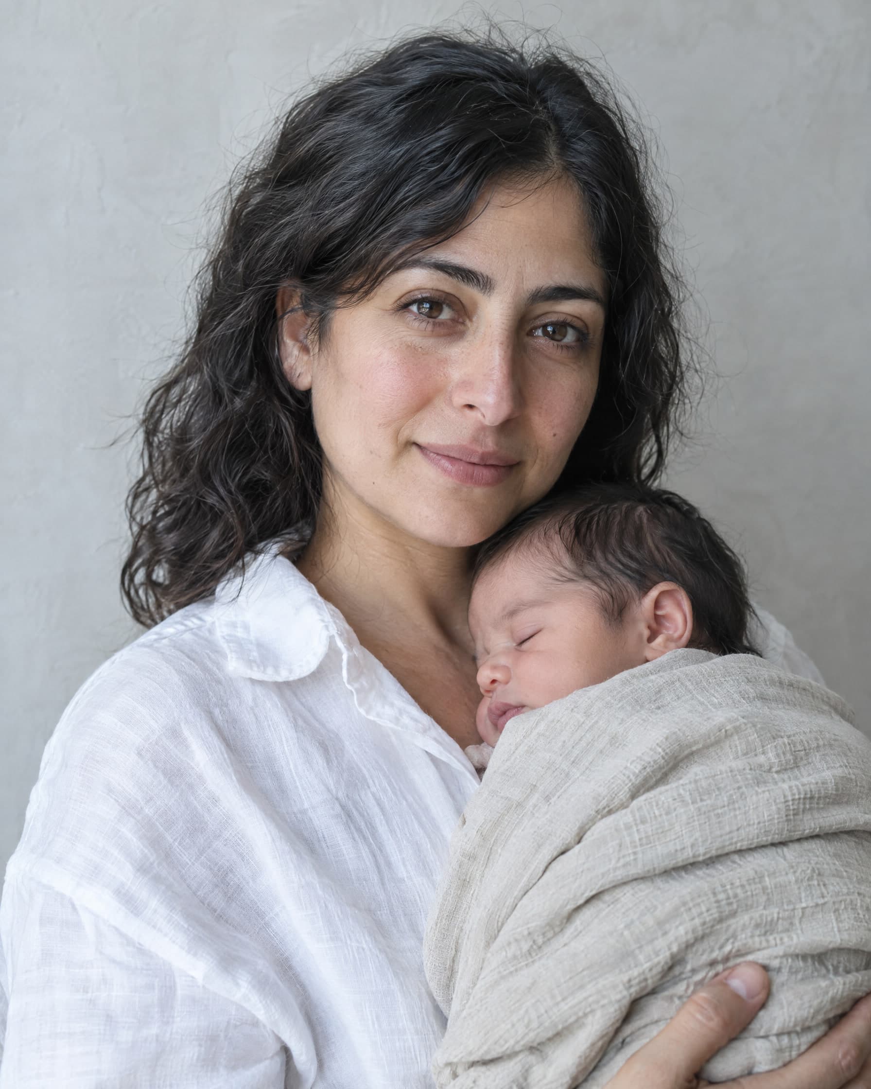 A woman in her mid-thirties with dark wavy hair, in a white linen shirt, holding a sleeping newborn wrapped in an oatmeal blanket against her chest.