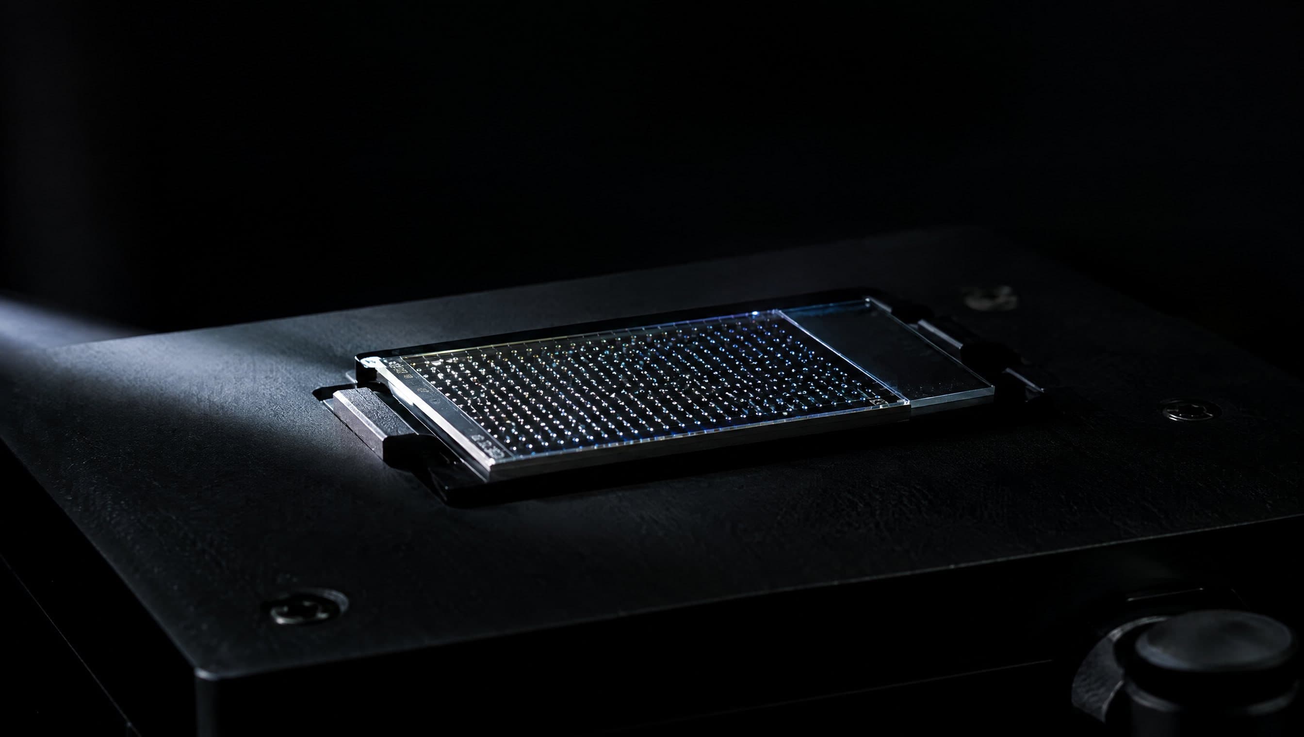 A clear plate of tiny sample spots on a dark instrument stage, lit by a narrow beam of light.