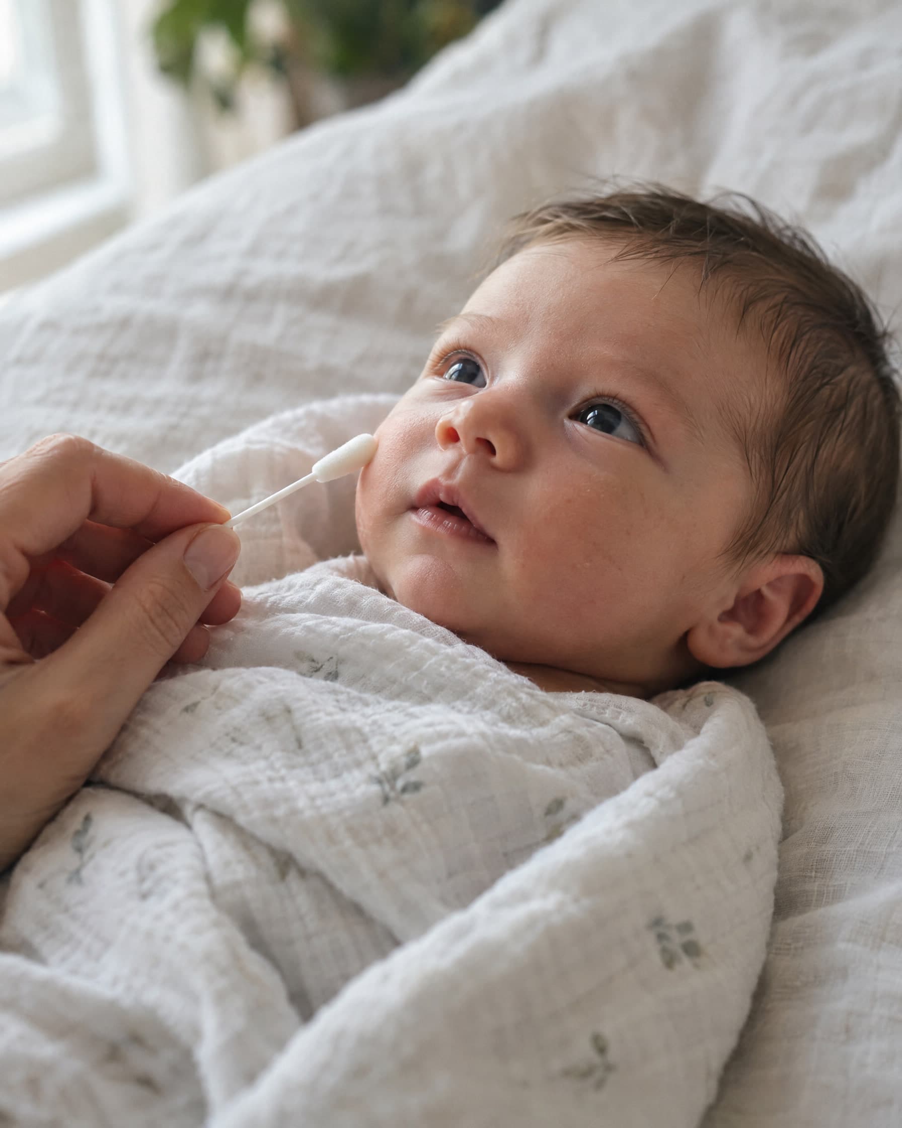 A hand holding a soft foam-tipped swab near a wide-eyed newborn swaddled in white muslin.