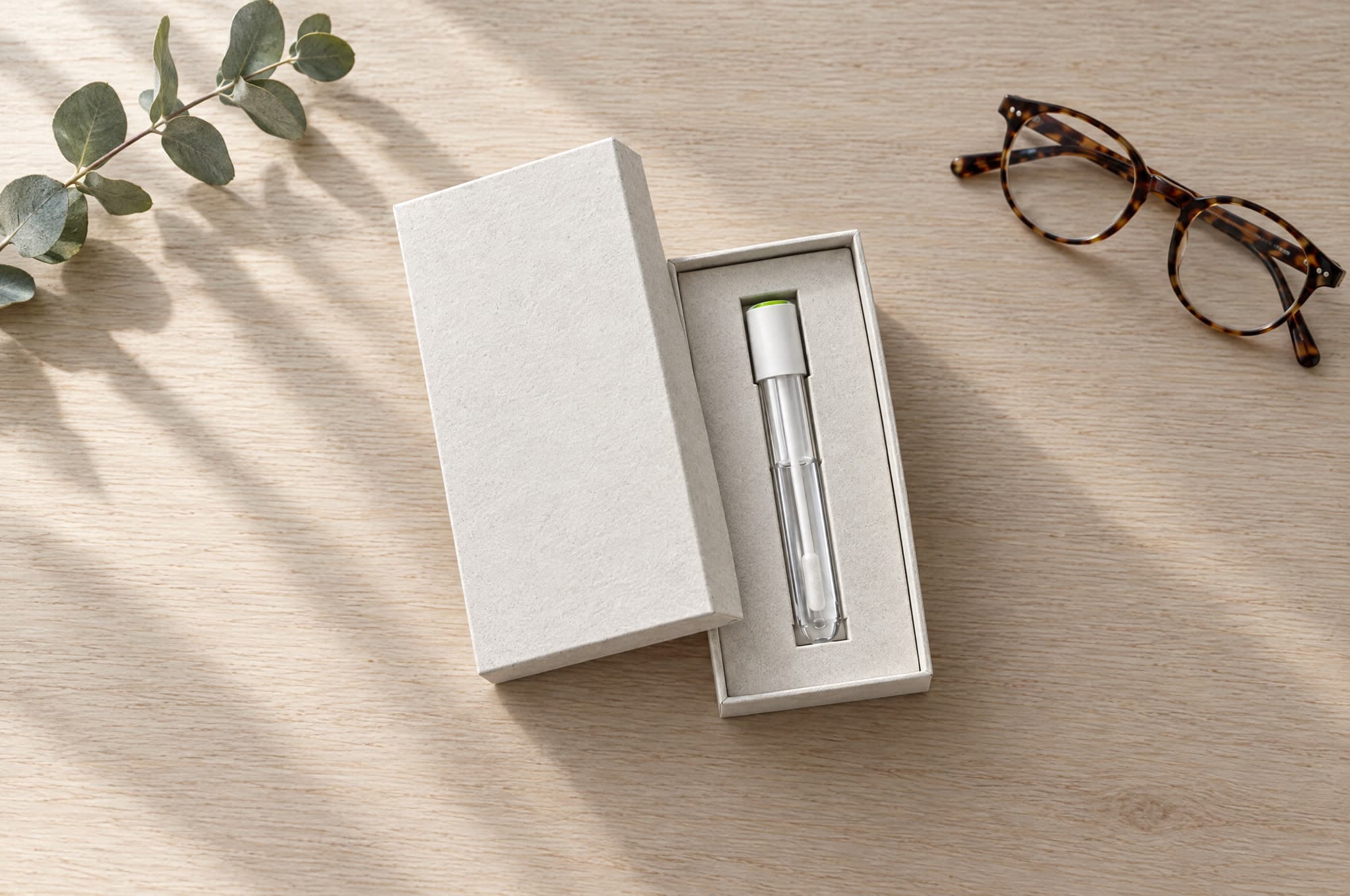 An open kit box with the collection tube inside, a eucalyptus sprig and a pair of glasses on the table beside it.