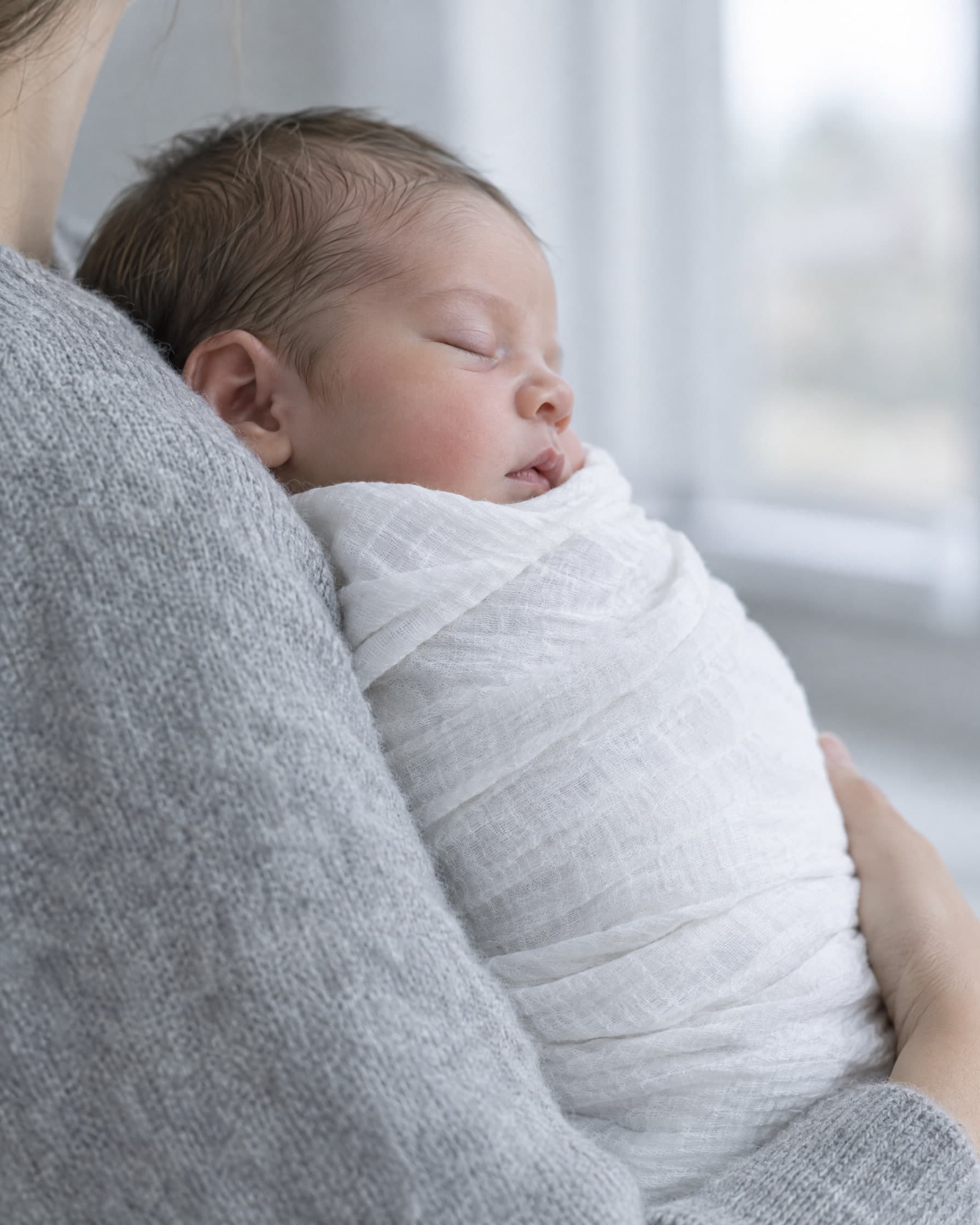 A parent holding a sleeping newborn, wrapped in white muslin, against their shoulder.