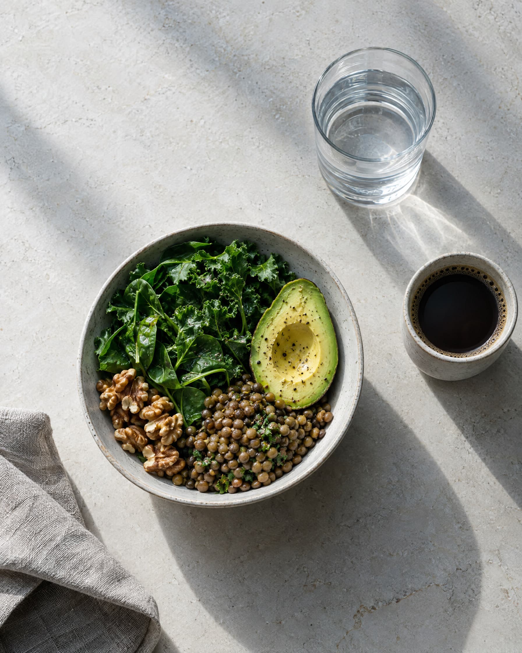From above: a bowl of greens, lentils, walnuts and half an avocado, with a glass of water and a black coffee.
