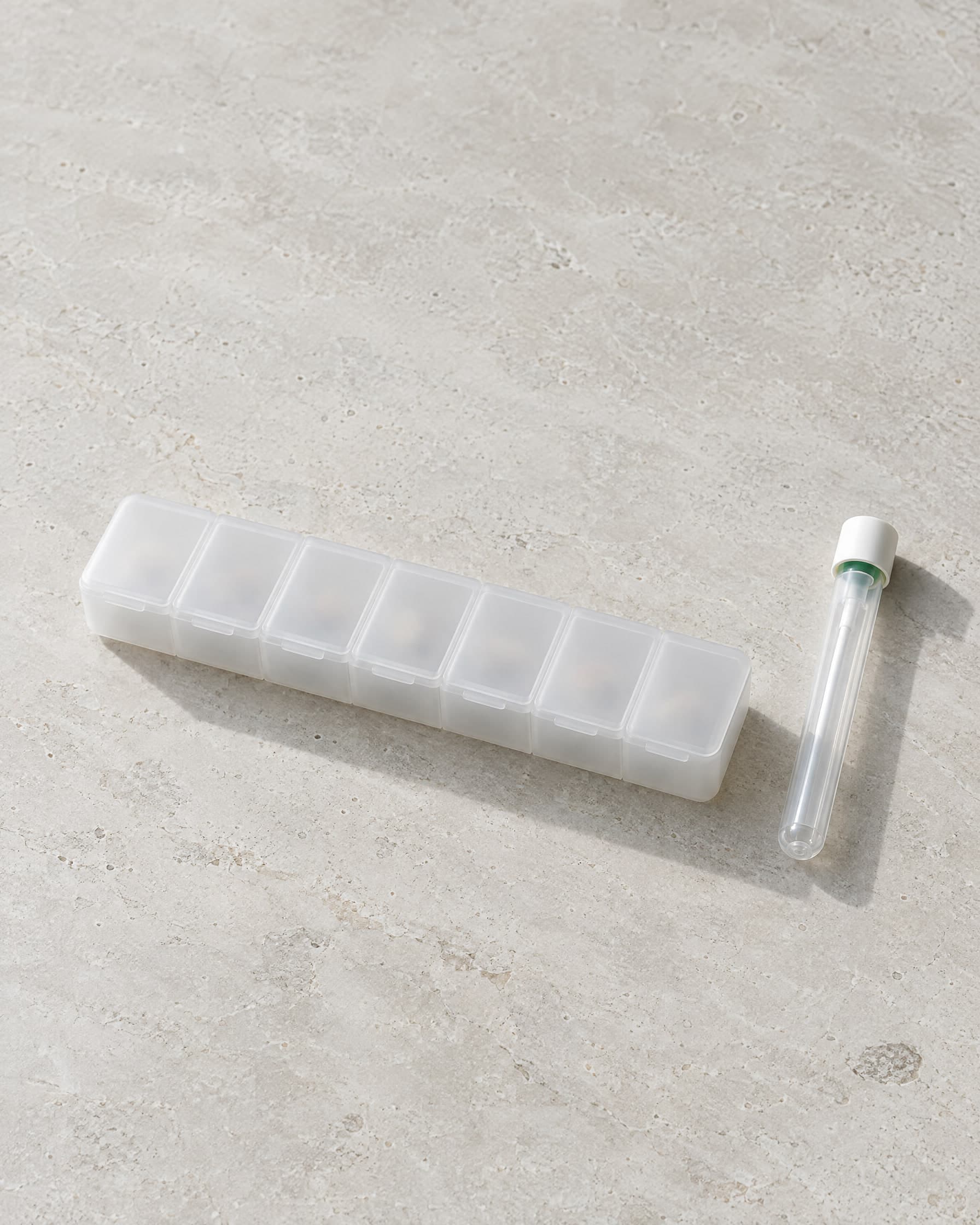 A frosted weekly pill organizer with blank lids beside the sample collection tube on pale stone.