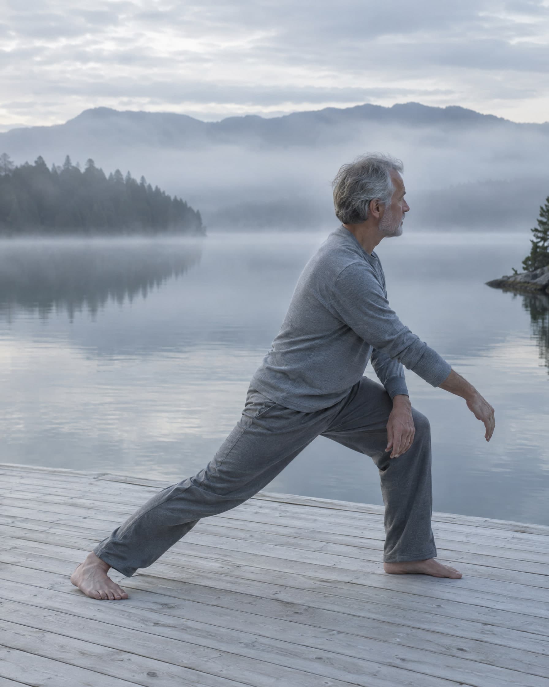 A man in his sixties stretching on a timber deck above a misty lake.